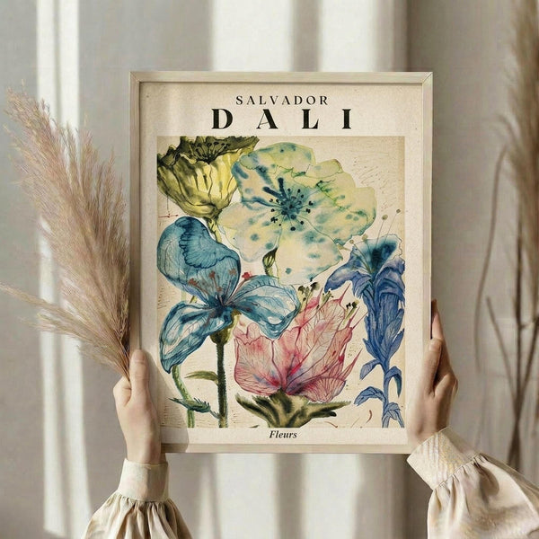 May include: Person in cream ruffled sleeves holding framed botanical print featuring watercolor flowers in blue, yellow, green and pink tones. Natural dried pampas grass visible on left side. Light wooden frame against soft neutral background with natural lighting. Vintage-style floral artwork with "Salvador Dali" and "Fleurs" text visible at top and bottom of print.