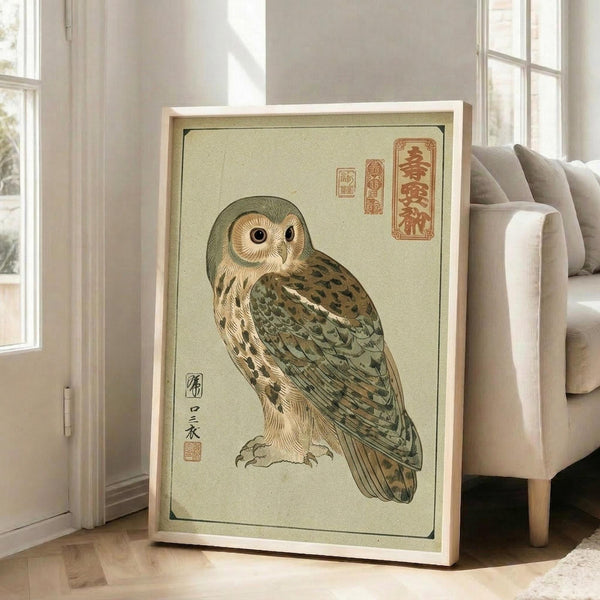 May include: Framed Japanese woodblock-style owl print in light wooden frame, leaning against white wall next to beige upholstered chair. The artwork features a detailed owl with mottled brown and green plumage on pale green background, with red Japanese calligraphy characters in upper right corner. Natural daylight streams through nearby windows onto hardwood floors in minimalist living room setting.