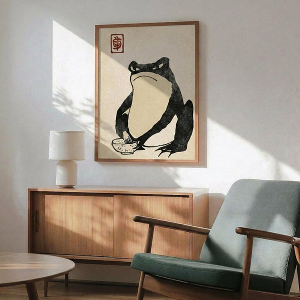 May include: Japanese-style artwork featuring a black silhouette frog in a contemplative pose holding a small bowl, displayed in a wooden frame above a mid-century modern walnut credenza. The minimalist print has a cream background with a red seal stamp. Scene shows teal upholstered chair with wood arms, white ceramic table lamp, and dramatic diagonal sunlight casting shadows across white walls.
