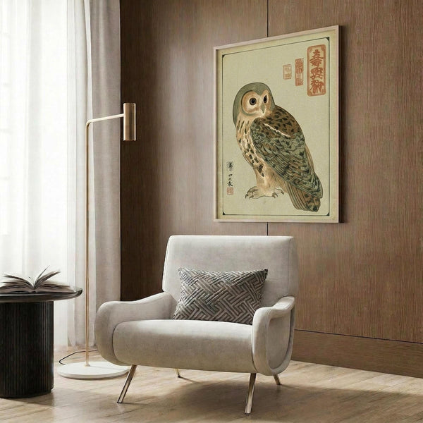 May include: A framed Japanese woodblock print of an owl with detailed brown and green feathers and Japanese calligraphy, displayed on a wood-paneled wall above a beige upholstered armchair with geometric patterned pillow. Scene includes a modern brass floor lamp and dark round side table with open book near a window with curtains.