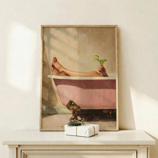 May include: Framed vintage-style artwork showing a person's legs and feet hanging over the edge of a pink clawfoot bathtub, holding a green margarita cocktail with lime garnish. The poster has warm, muted tones and is displayed in a light wood frame on a white shelf next to a small wrapped gift with greenery. Soft natural lighting creates gentle shadows on the cream-colored wall behind.