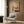 May include: Framed poster print hanging on wood-paneled wall above cream upholstered armchair with geometric pillow. Artwork shows woman's legs extending from pink vintage clawfoot bathtub while holding green cocktail. Modern living room setting with brass floor lamp, dark round side table with open book, and natural lighting from window with curtains.