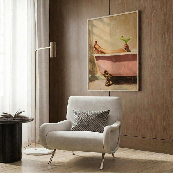 May include: Framed poster print hanging on wood-paneled wall above cream upholstered armchair with geometric pillow. Artwork shows woman's legs extending from pink vintage clawfoot bathtub while holding green cocktail. Modern living room setting with brass floor lamp, dark round side table with open book, and natural lighting from window with curtains.