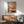 May include: Framed poster showing woman's legs in pink vintage bathtub holding green margarita with striped cat below, displayed above wooden sideboard with white lamp in modern living room with sage green chair and natural sunlight shadows on white wall.