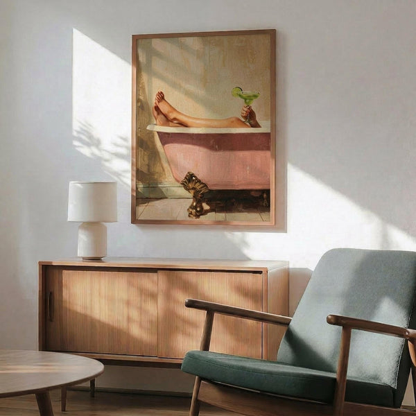 May include: Framed poster showing woman's legs in pink vintage bathtub holding green margarita with striped cat below, displayed above wooden sideboard with white lamp in modern living room with sage green chair and natural sunlight shadows on white wall.
