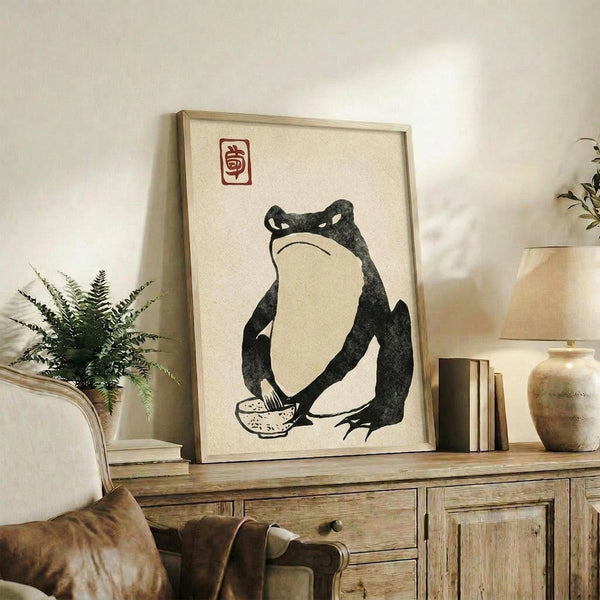 May include: Japanese-style artwork in light wood frame showing black ink frog holding small bowl, with red seal stamp above, displayed on rustic wooden sideboard beside fern plant, ceramic vase, and stacked books in neutral-toned living room setting