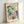 May include: A framed art print leaning against a white wall, featuring Salvador Dali's colorful floral illustration with blue, pink, yellow and green watercolor flowers on a cream background. The wooden frame sits on what appears to be a white surface, with soft natural lighting creating gentle shadows.