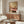 May include: Framed poster showing woman's legs in pink vintage bathtub holding green margarita cocktail, displayed above wooden mid-century credenza in modern living room with white curved chair, round glass coffee table, and white table lamp. Warm sunlight creates shadows across neutral walls and furniture.