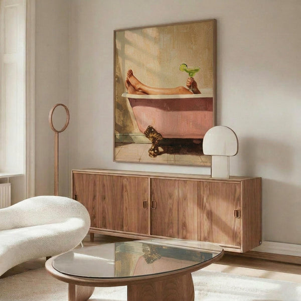 May include: Framed poster showing woman's legs in pink vintage bathtub holding green margarita cocktail, displayed above wooden mid-century credenza in modern living room with white curved chair, round glass coffee table, and white table lamp. Warm sunlight creates shadows across neutral walls and furniture.