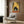 May include: Framed poster print displayed on wood paneled wall above cream upholstered armchair with geometric pillow. Artwork shows black cat with wide eyes eating noodles from bowl in Japanese-style kitchen setting with yellow tile walls, bottles, and warm lighting. Gold floor lamp and dark round table with open book visible in modern room scene.