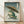 May include: Framed Japanese ukiyo-e style artwork showing a green frog holding a bamboo umbrella in the rain, displayed on light wooden flooring against a white wall. The print features traditional woodblock art styling with vertical rain lines, Japanese text characters, and muted green and beige tones in a natural wood frame.