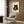 May include: Framed poster featuring a black cat with golden eyes eating sushi with chopsticks, displayed on wood-paneled wall above beige upholstered armchair with geometric patterned pillow. Modern brass floor lamp and dark side table with open book visible. Warm neutral interior with natural lighting from window.