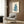 May include: Blue cat artwork in cream frame hanging on wood-paneled wall above beige upholstered armchair with geometric throw pillow. Modern reading corner with brass floor lamp, dark round side table with open book, and sheer curtains. The vintage-style blue cat illustration shows a sitting feline looking upward against cream background.