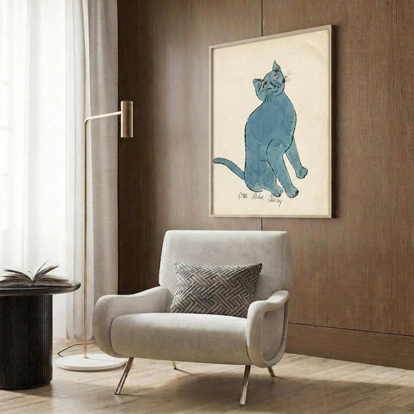 May include: Blue cat artwork in cream frame hanging on wood-paneled wall above beige upholstered armchair with geometric throw pillow. Modern reading corner with brass floor lamp, dark round side table with open book, and sheer curtains. The vintage-style blue cat illustration shows a sitting feline looking upward against cream background.