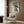 May include: Japanese woodblock print artwork featuring a green frog holding a bamboo umbrella in falling rain, displayed in light wooden frame on textured brown wall panel above cream upholstered armchair with geometric patterned pillow, alongside modern brass floor lamp and dark round side table with open book
