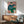 May include: Matisse portrait print in wooden frame hanging above mid-century modern sideboard with lamp, showing figure in blue shirt and green pants sitting on chair holding paper. Room has white walls, natural sunlight, teal upholstered armchair, and round coffee table creating contemporary living space mockup.