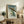 May include: Japanese woodblock print style artwork showing a green frog holding a bamboo umbrella in heavy rain, displayed in a light wooden frame on a rustic wooden sideboard. The print features traditional ukiyo-e aesthetics with vertical rain lines and Japanese text. Scene includes fern plant in white pot, ceramic vase, stacked books, and leather armchair in neutral-toned living room setting.