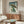 May include: Japanese ukiyo-e style artwork showing a green frog holding a bamboo umbrella in falling rain, displayed in a thin frame above a wooden sideboard. The print features traditional blue-green gradients with Japanese text characters. Modern living room setting with white curved chair, glass coffee table, floor lamp, and neutral walls with natural lighting.