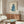 May include: Blue cat illustration artwork displayed in thin wooden frame on cream wall above mid-century walnut credenza. Modern living room scene with white curved chair, oval glass coffee table, and white dome table lamp. Cat sits upright with head tilted, rendered in watercolor style on beige background with cursive text below.