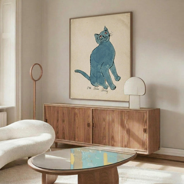 May include: Blue cat illustration artwork displayed in thin wooden frame on cream wall above mid-century walnut credenza. Modern living room scene with white curved chair, oval glass coffee table, and white dome table lamp. Cat sits upright with head tilted, rendered in watercolor style on beige background with cursive text below.