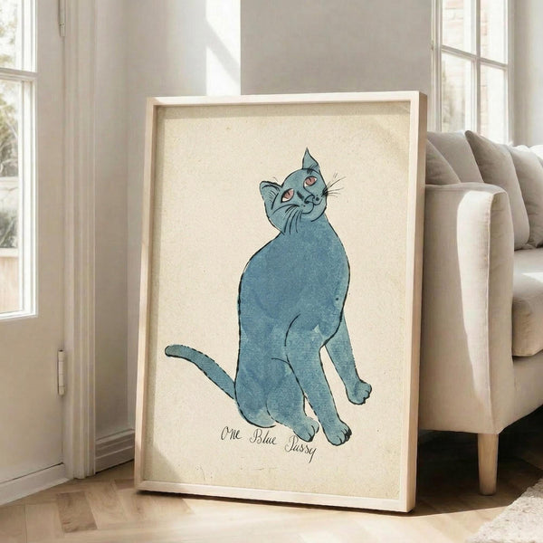 May include: Framed artwork featuring a blue cat sitting upright with head tilted back, rendered in watercolor style with orange eyes and whiskers. Text reads "One Blue Pussy" below the cat illustration. The piece is displayed in a light wood frame against a cream background, leaning against white walls near large windows in a bright living room with beige furniture.