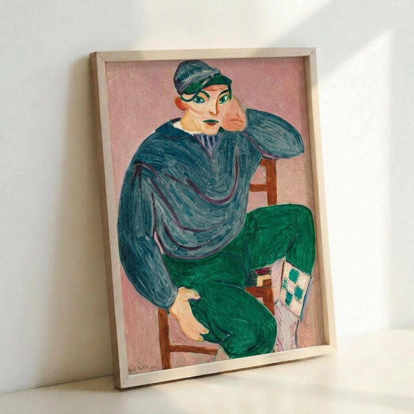 Framed reproduction of a Fauve-style painting showing a seated young person in a blue-green striped shirt and green pants, holding what appears to be a letter or paper. The figure wears a dark cap and has stylized facial features with bold colors including pink and green tones. The background is soft pink, and the painting is displayed in a light wooden frame leaning against a white wall, showing the artwork's vibrant brushstrokes and expressive color palette typical of early 20th century modern