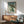 May include: Japanese woodblock-style artwork showing a green frog holding an umbrella in falling rain, displayed in a wooden frame above a mid-century modern credenza. The print features traditional ukiyo-e art style with Japanese characters, mounted on a white wall next to a ceramic table lamp and sage green upholstered chair with wooden arms.