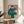 May include: Matisse-style portrait print in thin wooden frame displayed on round wooden side table next to beige upholstered chair. Artwork shows figure in teal blue cap and sweater with green pants, sitting in contemplative pose against pink background. Modern minimalist room setting with white walls, natural lighting, and plant leaves visible in corner.