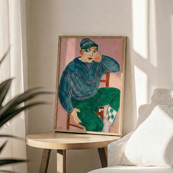 May include: Matisse-style portrait print in thin wooden frame displayed on round wooden side table next to beige upholstered chair. Artwork shows figure in teal blue cap and sweater with green pants, sitting in contemplative pose against pink background. Modern minimalist room setting with white walls, natural lighting, and plant leaves visible in corner.