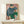 May include: Framed art print displaying a seated figure in blue top and green pants against pink background, shown in modern interior setting on light wood console table with small white wrapped gift, natural lighting creating soft shadows on cream wall