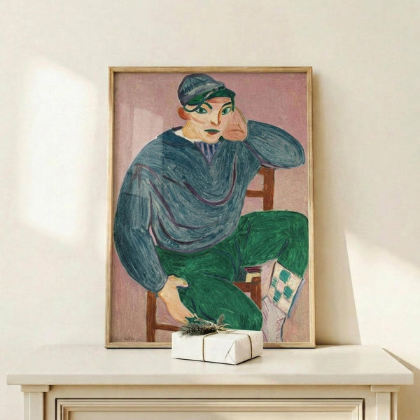 May include: Framed art print displaying a seated figure in blue top and green pants against pink background, shown in modern interior setting on light wood console table with small white wrapped gift, natural lighting creating soft shadows on cream wall