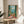 May include: Framed artwork displaying a Mediterranean balcony scene with turquoise shutters, wicker chair, potted flowers, and ocean view, positioned on a round wooden side table next to a beige upholstered chair in a sunlit room with white walls and plant shadows