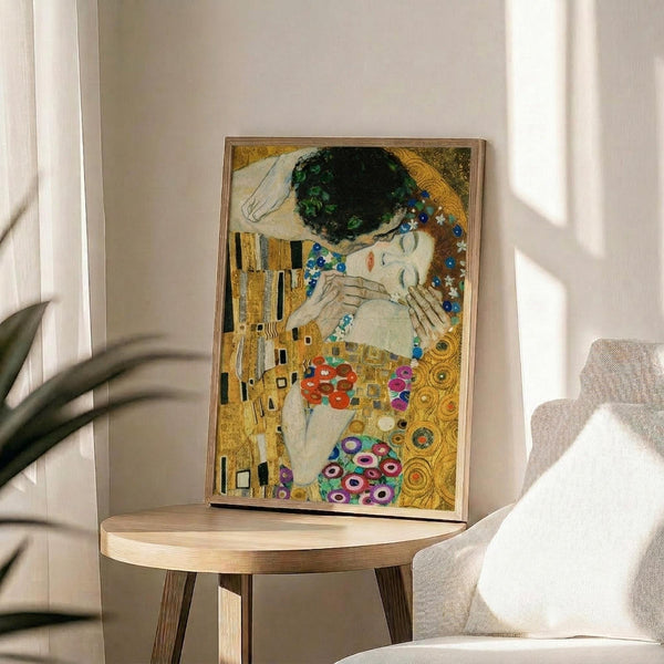 May include: Framed reproduction of Gustav Klimt's famous painting showing two embracing figures in ornate golden robes with geometric patterns and colorful circular motifs, displayed on a round wooden side table next to a beige upholstered chair with white pillow, green plant leaves visible on left, natural sunlight streaming across white walls creating soft shadows