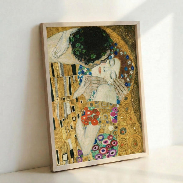 May include: Framed reproduction of Klimt's famous painting showing an embracing couple against ornate golden background with decorative patterns, circles, and geometric designs. The woman has closed eyes and dark hair, wearing a colorful floral dress. The artwork features rich golds, blues, greens, and jewel tones in Klimt's distinctive Art Nouveau style. Light wooden frame positioned against white wall.