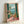 A framed art print leaning against a white wall showing a Mediterranean balcony scene with green shutters, wicker chair, potted plants with pink flowers, and a coastal view with palm trees and blue water. The artwork features warm coral and turquoise tones with a vintage 1950s illustration style. The print is displayed in a light wood frame on what appears to be a white surface.