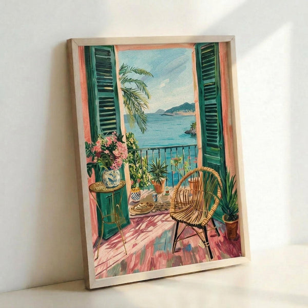 A framed art print leaning against a white wall showing a Mediterranean balcony scene with green shutters, wicker chair, potted plants with pink flowers, and a coastal view with palm trees and blue water. The artwork features warm coral and turquoise tones with a vintage 1950s illustration style. The print is displayed in a light wood frame on what appears to be a white surface.