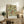 May include: Framed botanical poster with intricate floral pattern in greens, golds, and coral red displayed on rustic wooden sideboard. Room features neutral walls, fern plant, ceramic vase, stacked books, and brown leather chair arm visible in foreground.