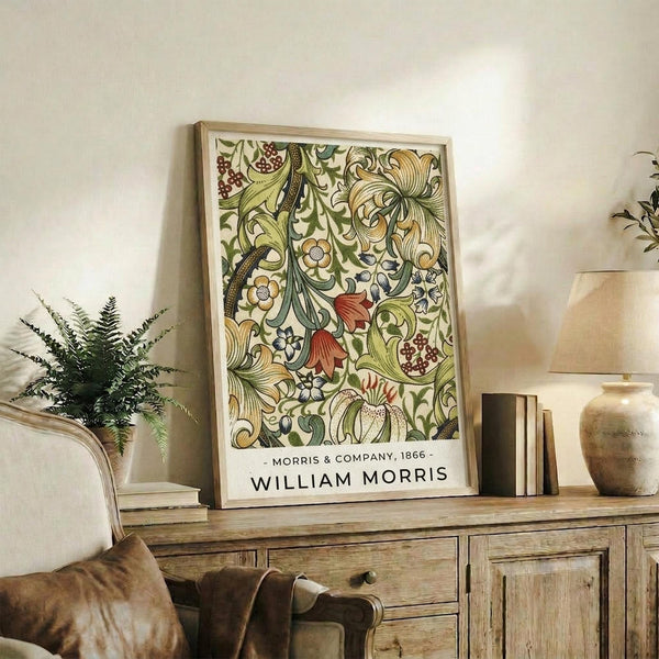 May include: Framed botanical poster with intricate floral pattern in greens, golds, and coral red displayed on rustic wooden sideboard. Room features neutral walls, fern plant, ceramic vase, stacked books, and brown leather chair arm visible in foreground.