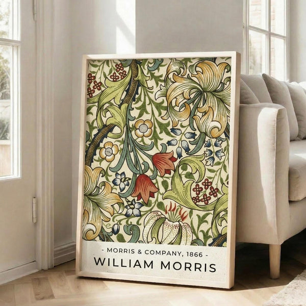 May include: Framed botanical art print with intricate floral pattern featuring green leaves, golden scrollwork, red berries, and cream flowers in William Morris Victorian style, displayed against white wall next to beige upholstered chair in bright living room with large windows and natural light streaming across wooden floors.