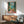 May include: Framed vintage-style poster depicting a Mediterranean balcony scene with green shutters, wicker chair, potted plants, and ocean view, displayed above a mid-century modern wooden credenza with table lamp in a contemporary living room setting with teal upholstered chair and natural sunlight casting shadows on white walls.