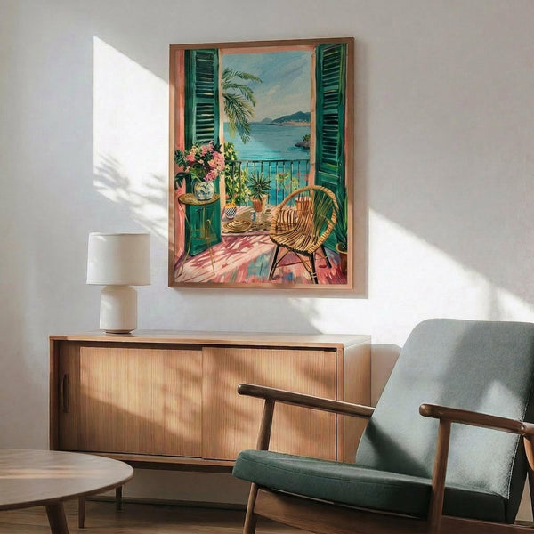 May include: Framed vintage-style poster depicting a Mediterranean balcony scene with green shutters, wicker chair, potted plants, and ocean view, displayed above a mid-century modern wooden credenza with table lamp in a contemporary living room setting with teal upholstered chair and natural sunlight casting shadows on white walls.