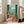 May include: Large framed vintage-style poster showing Mediterranean balcony scene with wicker chair, coral walls, green shutters, ocean view with mountains, decorative vase with pink flowers, palm fronds, and geometric tiled floor. Print displayed in bright white living room next to beige sofa and tall windows.