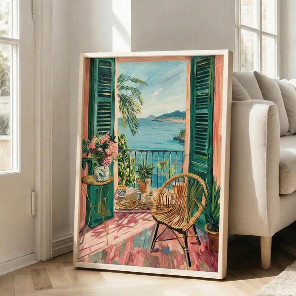May include: Large framed vintage-style poster showing Mediterranean balcony scene with wicker chair, coral walls, green shutters, ocean view with mountains, decorative vase with pink flowers, palm fronds, and geometric tiled floor. Print displayed in bright white living room next to beige sofa and tall windows.