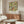 May include: Framed botanical art print featuring intricate green foliage, red berries, and cream flowers in Victorian decorative style, displayed above a walnut wood credenza with brass handles. Modern white swivel chair and glass-top coffee table visible in minimalist living room with beige walls and natural lighting.