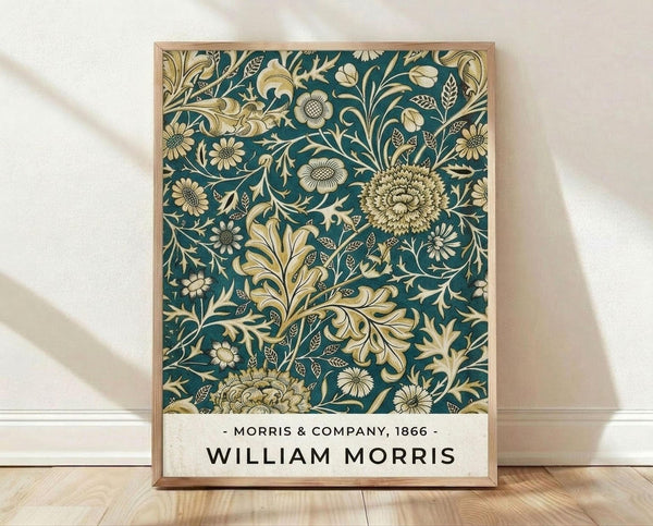 May include: Framed art print featuring an intricate Victorian floral pattern with golden chrysanthemums, ornate leaves, and decorative flowers on a deep teal background. The vintage-style botanical design fills most of the frame, with "Morris & Company, 1866" and "William Morris" text at the bottom on a cream banner. The wooden frame leans against white fabric draped behind it, sitting on light wood flooring with natural shadows cast across the scene.