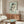 May include: Framed art print of a curled nude figure in earth tones against pale green background, displayed above mid-century walnut credenza in modern living room with cream chair, brass floor lamp, oval glass coffee table, and white walls with natural sunlight