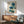 May include: Framed botanical artwork featuring blue poppies with Asian calligraphy characters displayed above a wood sideboard with table lamp, next to a sage green mid-century armchair in a bright living room with white walls and natural sunlight streaming across the scene.