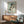 May include: Framed art print of a crouching female figure in muted earth tones and sage green background, displayed above a mid-century modern walnut credenza with table lamp. Green upholstered armchair with wooden frame sits beside round wood coffee table in bright, naturally-lit room with white walls and dramatic window shadows.