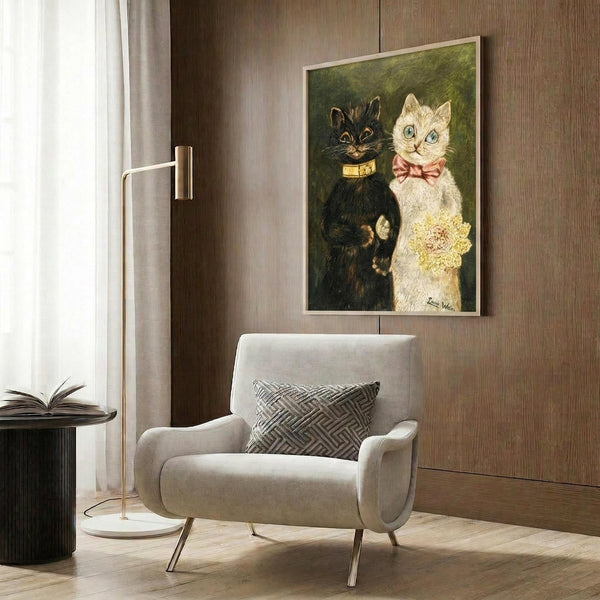 May include: Framed vintage-style cat wedding portrait showing two cats in formal attire - a black cat in golden collar and white cat in pink bow tie holding yellow flowers. Art hangs on wood-paneled wall above beige upholstered armchair with geometric pillow. Modern floor lamp and side table with open book visible in warm-toned room setting.