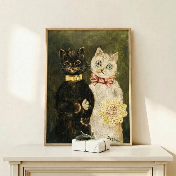 May include: Framed vintage-style cat wedding illustration displayed on white wooden mantel. Two anthropomorphic cats in formal attire - black cat with gold collar beside white cat with pink bow tie holding yellow flowers. Dark green textured background. Small white gift box with sprig sits on mantel. Warm lighting creates soft shadows on cream wall.